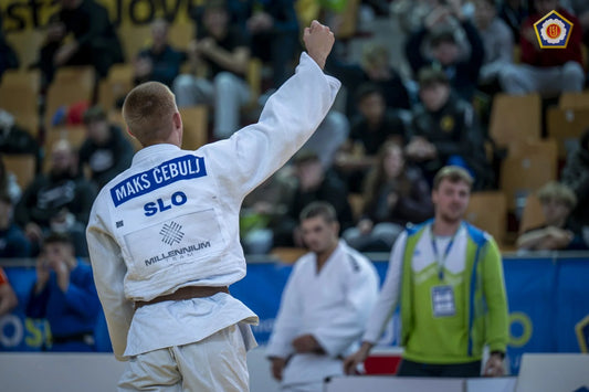 Judoka in a white gi raises a clenched fist toward the crowd, with a back patch reading Maks Čebulj and SLO.