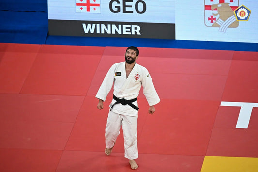 A judoka in a white gi and black belt stands barefoot on a red tatami beneath a screen showing GEO and WINNER.