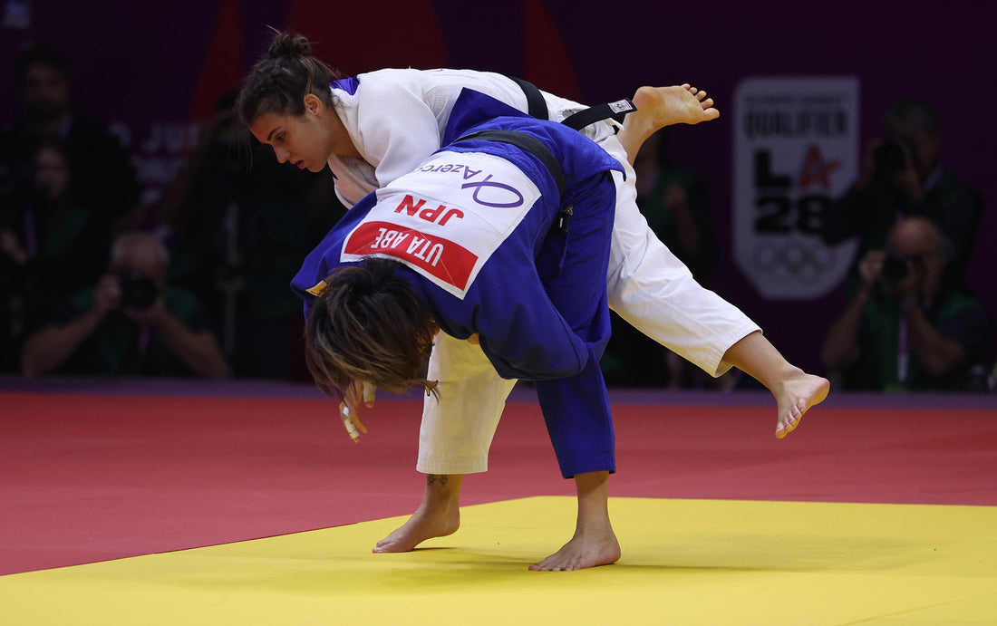 A judoka in a white judogi lifts an opponent in a blue judogi during a standing exchange on a yellow and red tatami.