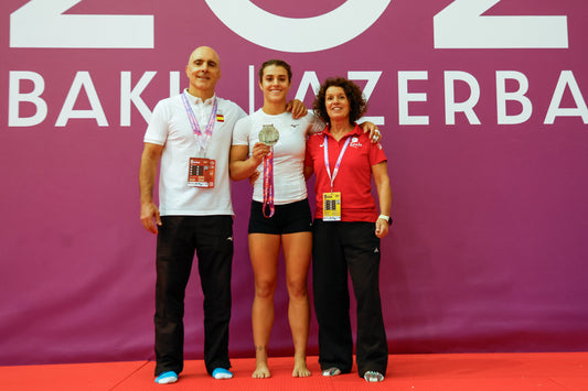 Three people stand barefoot or in trainers on a red mat in front of a large Baku Azerbaijan backdrop, with the central athlete holding a medal.