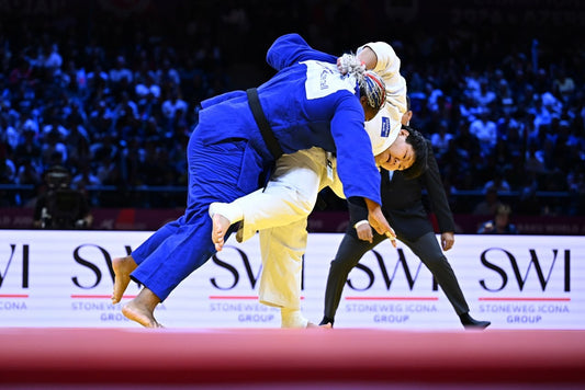A judoka in white lifts a judoka in blue during a throw, while a referee watches on the competition mat.