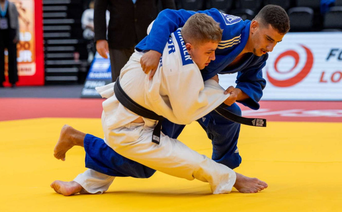 Two judoka in white and blue judogi grapple on a yellow tatami during a match.