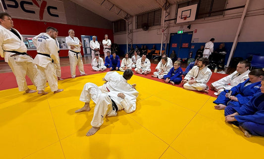 Judoka in white and blue judogi sit and stand around a yellow tatami while two athletes demonstrate on the mat.