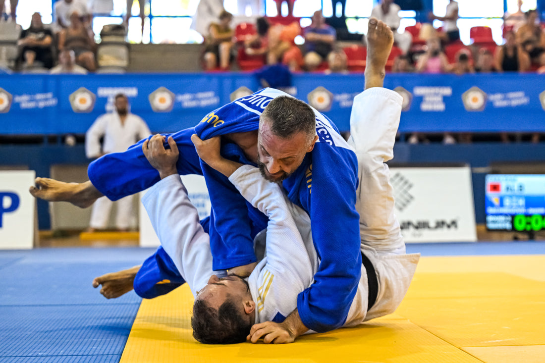 A competitor in a blue judogi grapples with an athlete in a white judogi on a yellow and blue tatami, with spectators in the background.