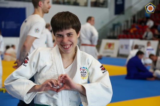 A smiling judoka in a white judogi makes a heart shape with their hands on a blue and yellow tatami.