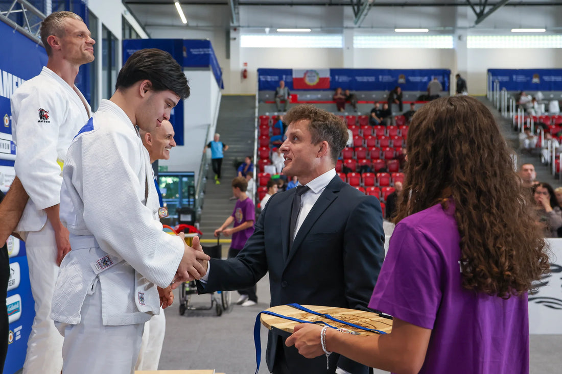 A man in a dark suit shakes hands with a judoka in a white judogi beside a podium, while another person holds medals.