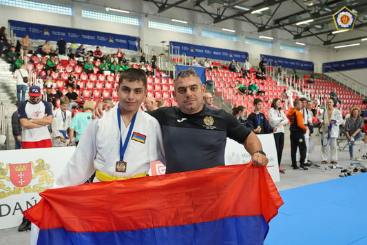A young judoka in a white judogi and yellow belt stands with a man while they hold an Armenian flag; the judoka wears a medal.
