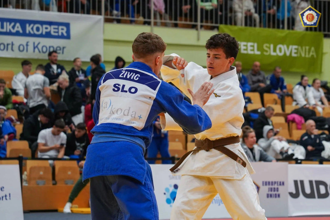 Two judoka in blue and white judogi grip each other during a match, with spectators seated in the background.
