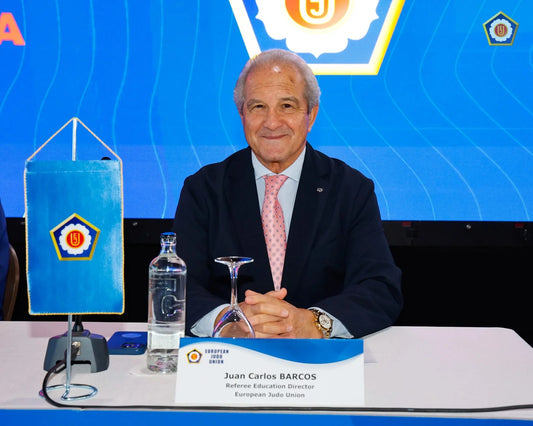 Juan Carlos Barcos sits at a table in a dark suit and pink tie, with a nameplate, water bottle and European Judo Union banner.