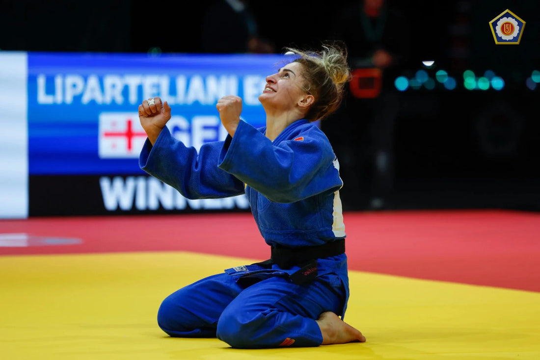Judoka in a blue gi kneeling on the yellow tatami with raised fists, with a competition screen in the background.