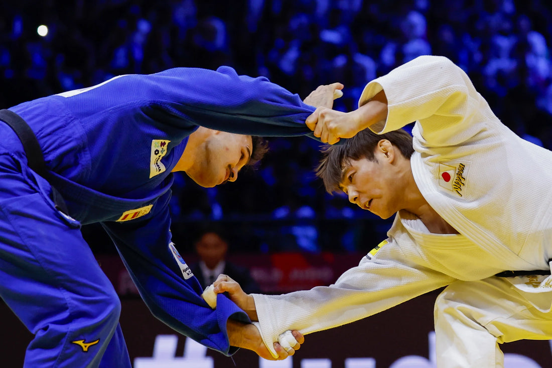 A judoka in a blue judogi and an opponent in white grip each other’s sleeves during a bout.