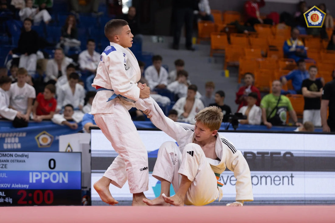 Two judoka in white judogi during a match, with one standing and holding the other’s sleeve as spectators watch from the stands.