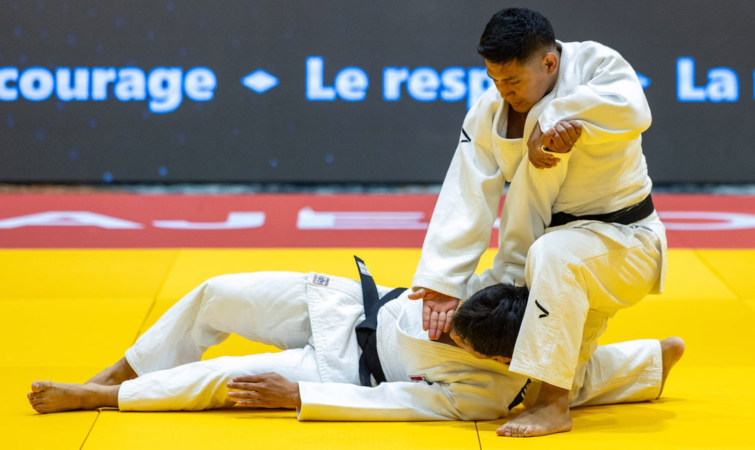 Two judoka in white uniforms on a yellow mat, with one holding the other as they lie on the tatami.