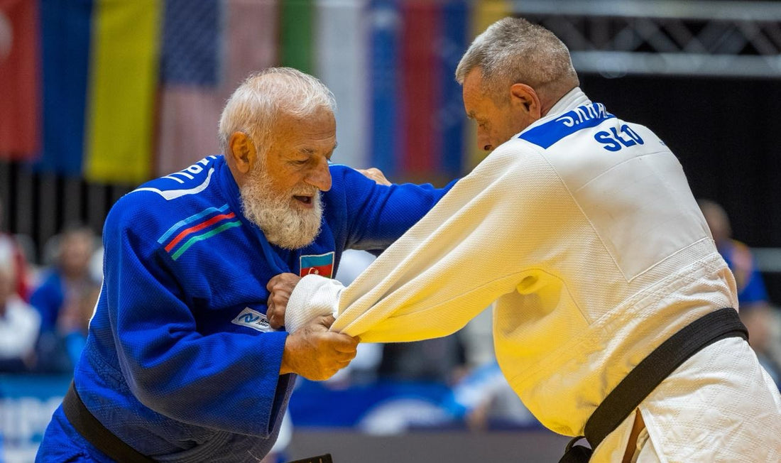 Two older judoka in blue and white Judogi grip each other during a standing exchange on the tatami.