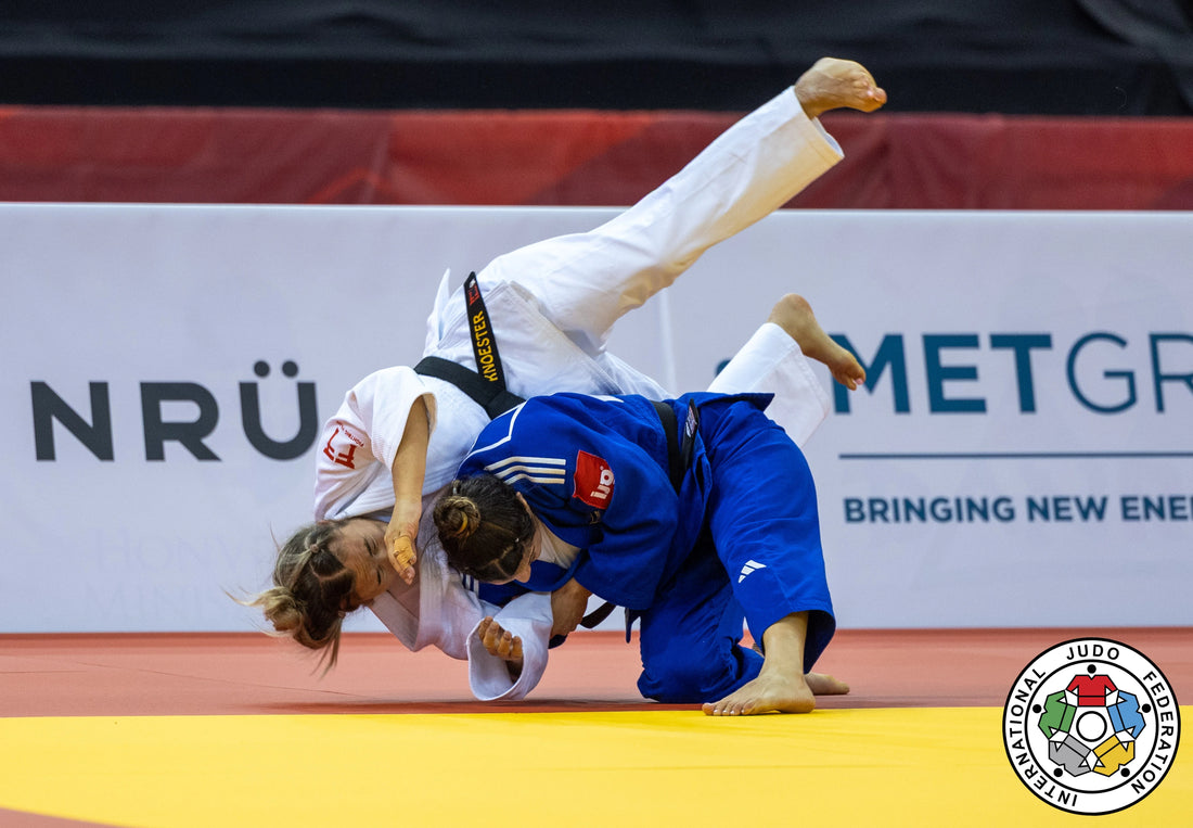 A judoka in a white judogi is lifted and tilted over an opponent in a blue judogi on a yellow tatami.