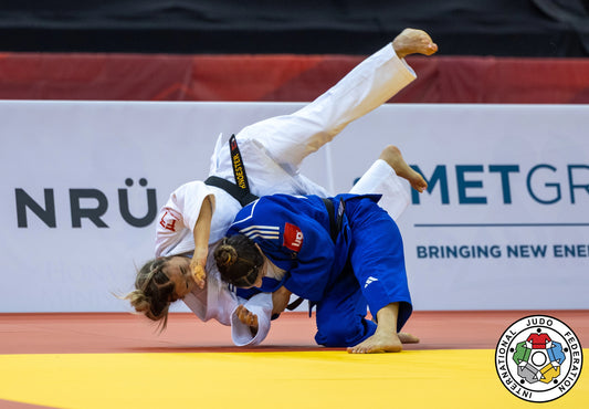 A judoka in a white judogi is lifted and tilted over an opponent in a blue judogi on a yellow tatami.