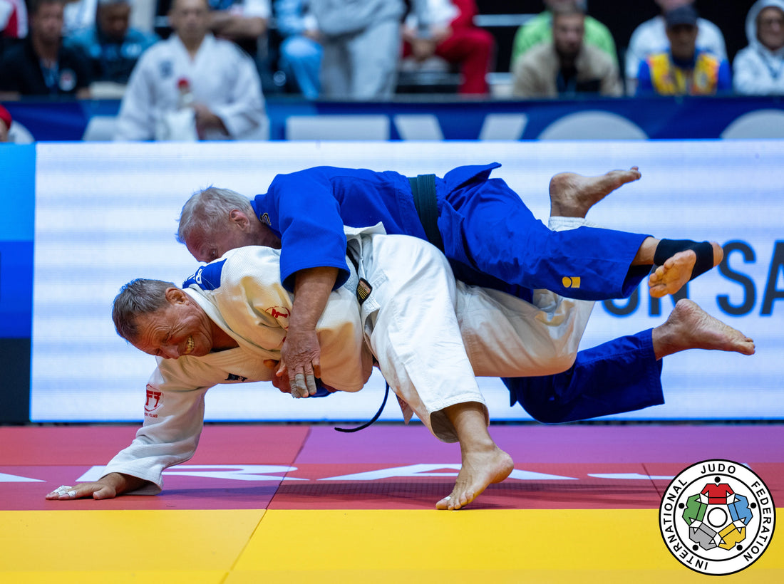 Two judoka in blue and white judogi grapple during a throw on a yellow and red tatami, with spectators behind them.