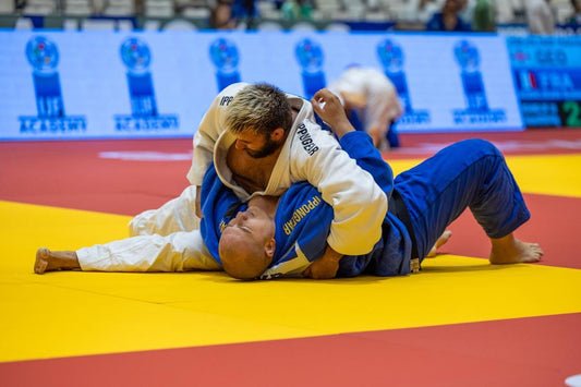 Two judoka in white and blue judogi grapple on the yellow tatami during a ground exchange, with scoreboards visible behind them.