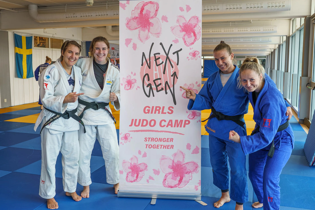 Four women in white and blue judogi pose beside a Next Gen Girls Judo Camp banner on blue and yellow tatami mats.